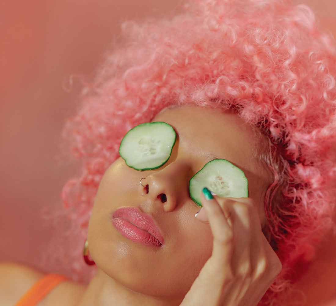 A woman with curly pink heir laying down with cucumbers over her eyes